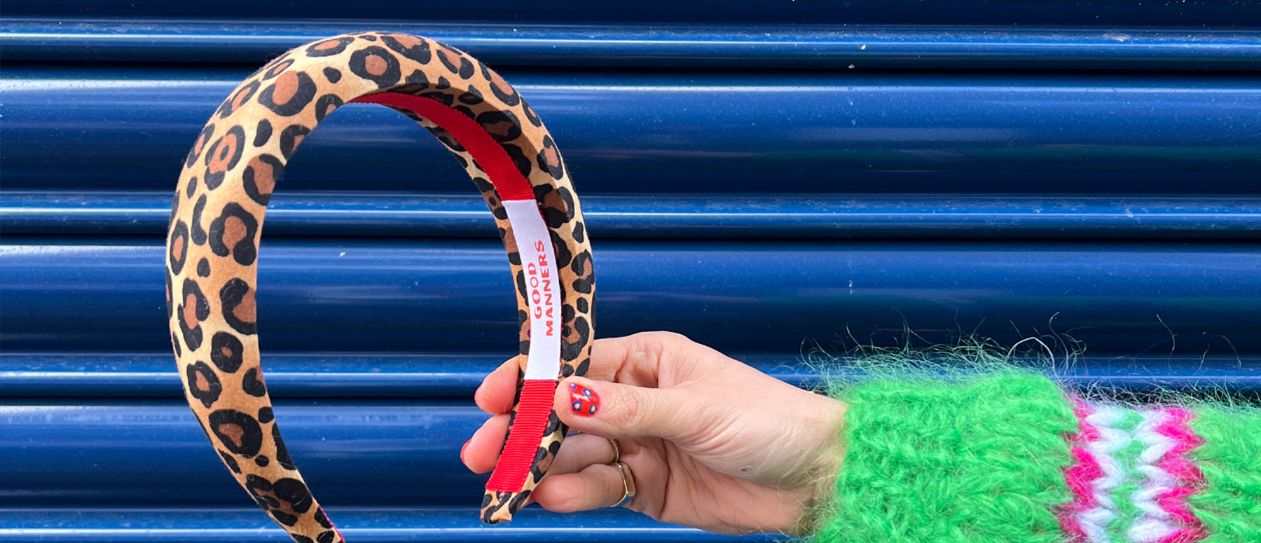 good manners style leopard headband held up in front of blue shutters hand has red manicure